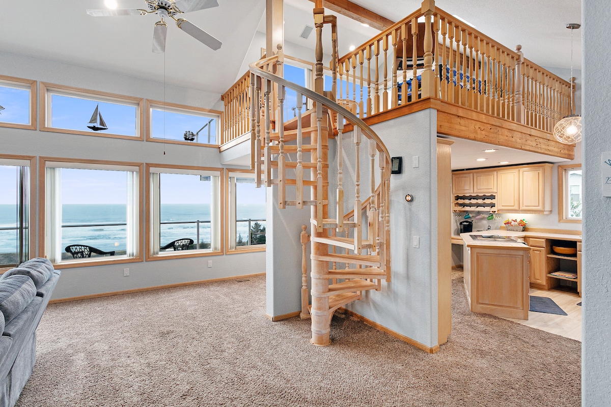 A spacious open floor plan is showcased, featuring large windows with panoramic ocean views. A wooden spiral staircase leads to the upper level. The living area includes carpeted flooring and a comfortable seating arrangement, creating an inviting space filled with natural light.