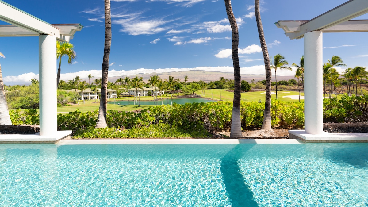 A private infinity pool reflects the clear blue sky and lush greenery of the golf course beyond. Towering palm trees frame the scene, enhancing the serene landscape, while distant hills create a tranquil backdrop, inviting relaxation and enjoyment of the stunning views.
