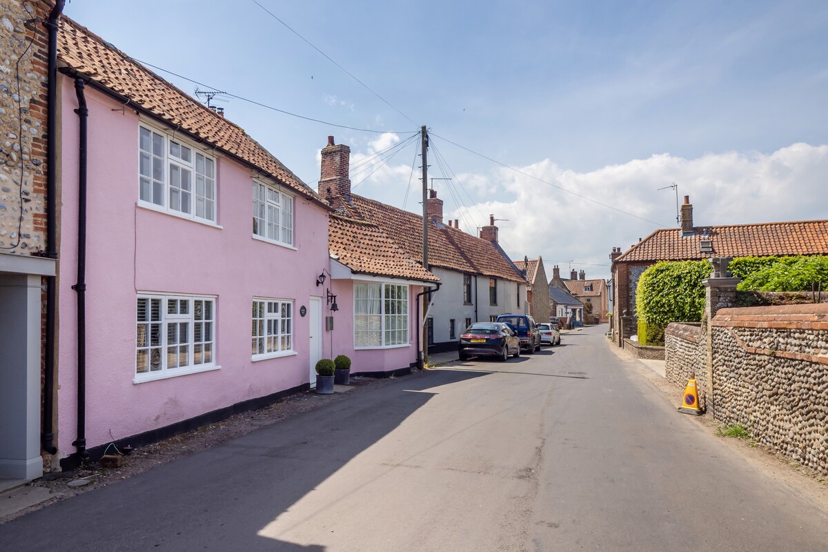 A charming street view captures a line of artisan cottages featuring traditional flint stone and painted facades. Notable is the pink cottage on the corner, adorned with white window frames. The tidy street is lined with parked vehicles and edged by green hedges, creating a quaint village atmosphere.