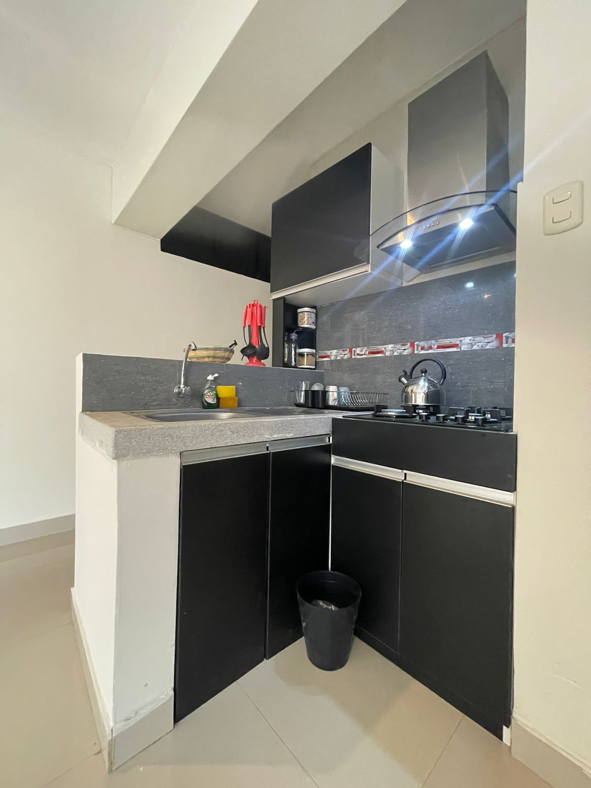 The compact kitchen features sleek black cabinetry and a gray countertop. Essential appliances, including a stove and coffee maker, are visible, while utensils are neatly arranged. Soft lighting enhances the functional space, creating an efficient and organized cooking area.