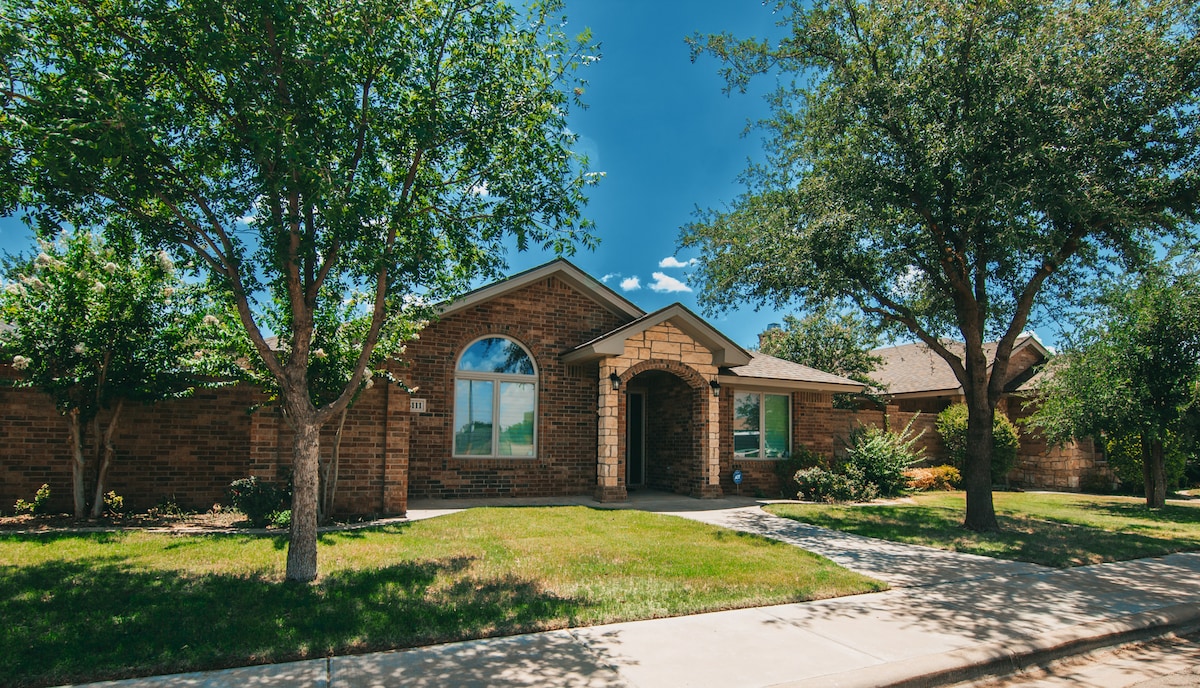 A charming single-story brick home is surrounded by green lawns and trees. A welcoming arched entrance is framed by two large windows, allowing natural light to brighten the interior. The walkway leads to the front door, inviting guests into a serene environment.