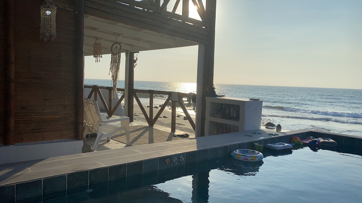 A sunlit scene presents a pool adjacent to a spacious deck, framed by wooden beams. Gentle waves are seen lapping against the shore in the background, while various pool toys float on the water's surface. The sky features soft hues of gold and blue.