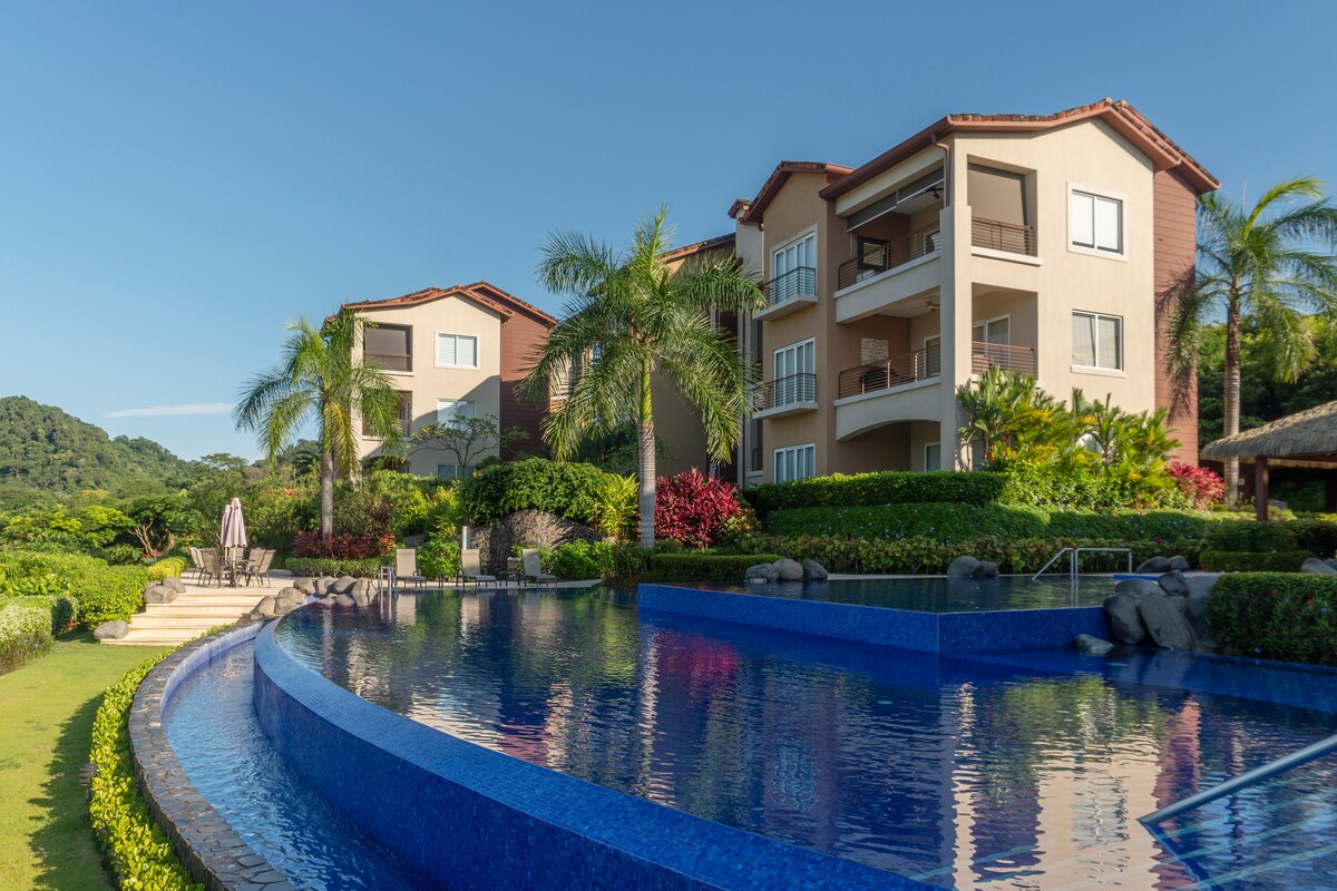 An expansive view of the resort showcases a blue infinity pool bordered by lush greenery and palm trees. The three-story building offers balconies with a scenic backdrop of vibrant landscaping and distant hills, creating a serene outdoor environment.