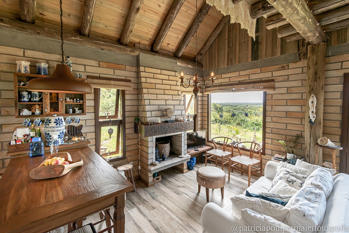 A cozy living area features wooden beams and a stone fireplace. Large windows offer expansive views of the surrounding landscape. A wooden dining table and chairs are positioned adjacent to the kitchen. Decorative items are placed on shelves, enhancing the inviting ambiance.