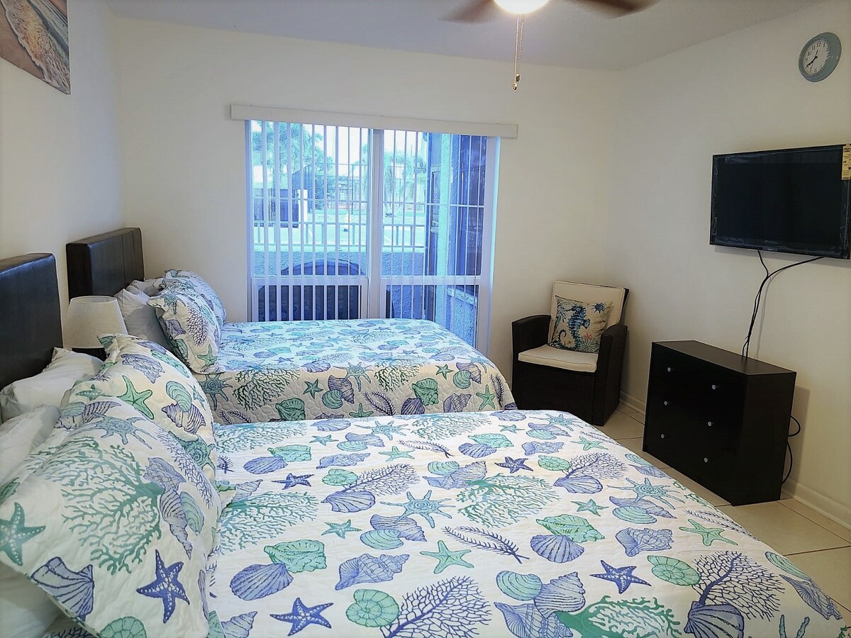 A bright and airy bedroom features two spacious beds adorned with a marine-themed bedding set. Natural light enters through sliding glass doors with blinds, illuminating a comfortable seating area with a small chair. A flat-screen TV is mounted on the wall opposite a wooden dresser.