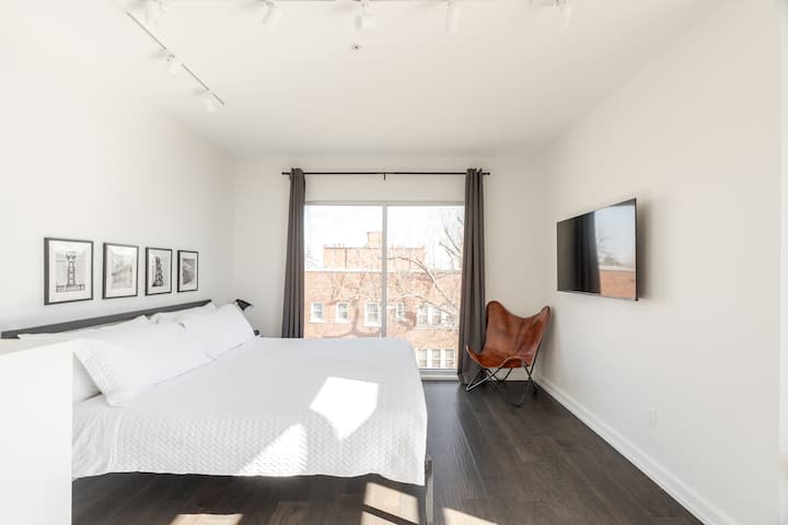 Bright, minimalist bedroom with crisp white linens, framed art, and a cozy leather chair. Natural light and modern touches create a peaceful urban retreat.