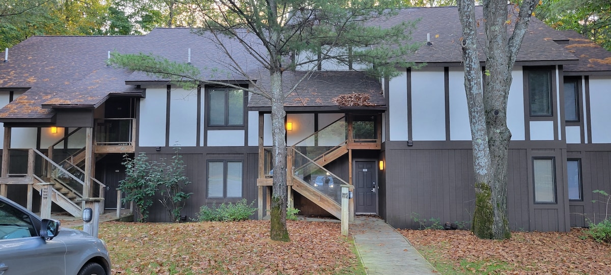A two-story condo building is surrounded by a wooded setting, with autumn leaves covering the ground. The exterior features a mix of dark wood and light-colored panels, complemented by large windows. Stairs lead to the entrance, illuminated by subtle outdoor lighting.