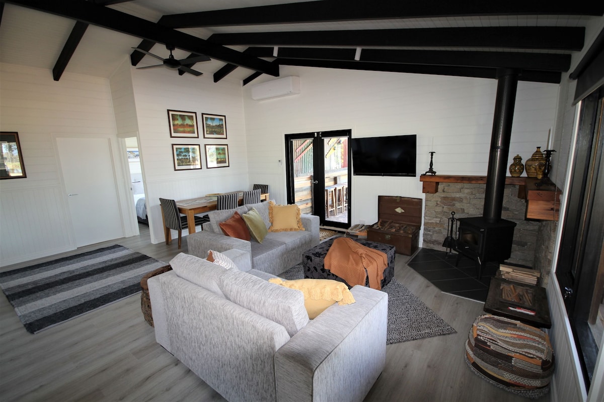 A spacious living area features a modern design with high ceilings and exposed beams. Two comfortable sofas are positioned on a textured rug. A rustic stone fireplace complements the seating, while a dining area and large glass doors are visible in the background.