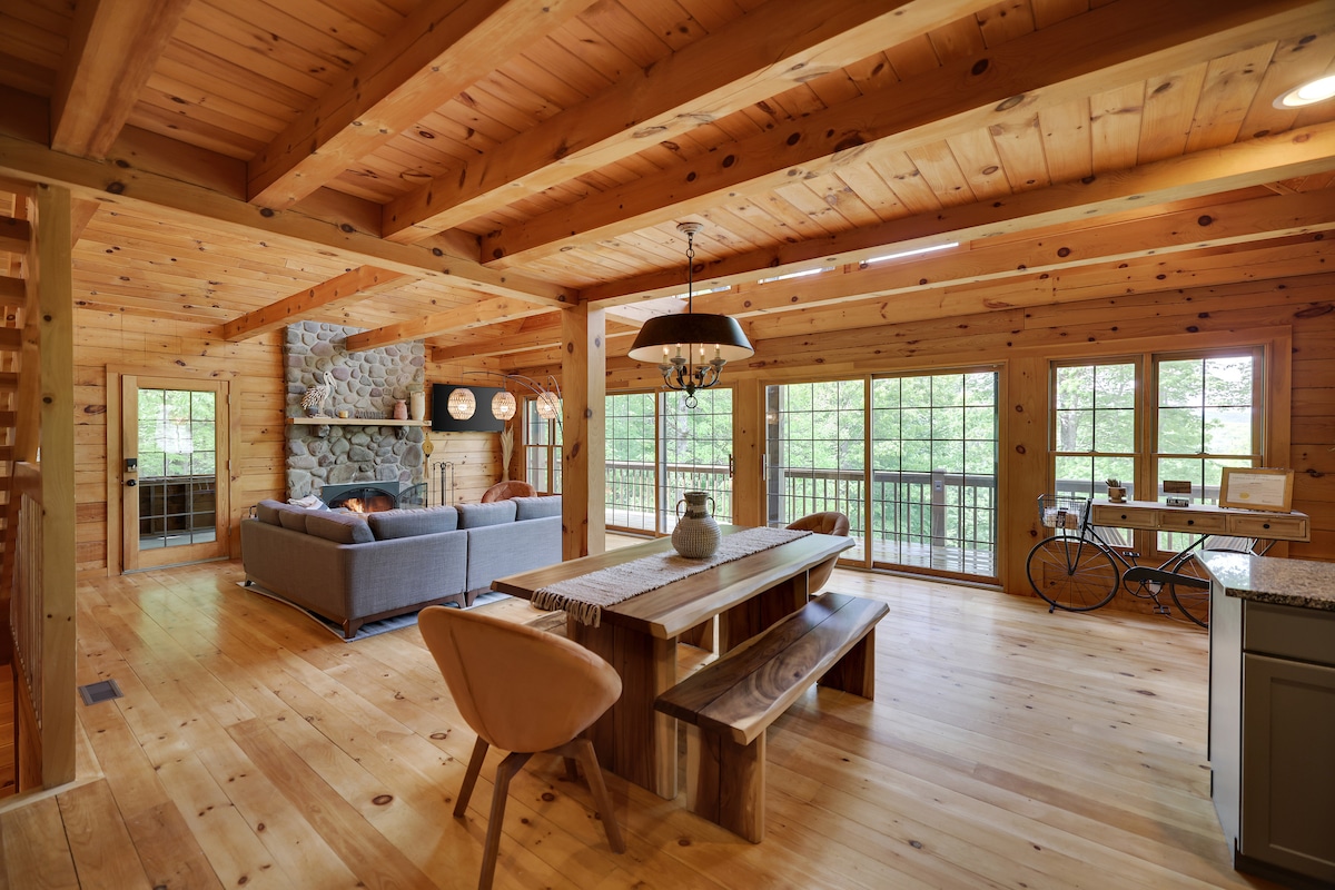 An open dining and living area features a large wooden table with unique seating and a cozy gray sofa. Natural light filters through expansive windows, illuminating the wooden beams and floors. A stone fireplace serves as a focal point, adding warmth to the rustic interior.