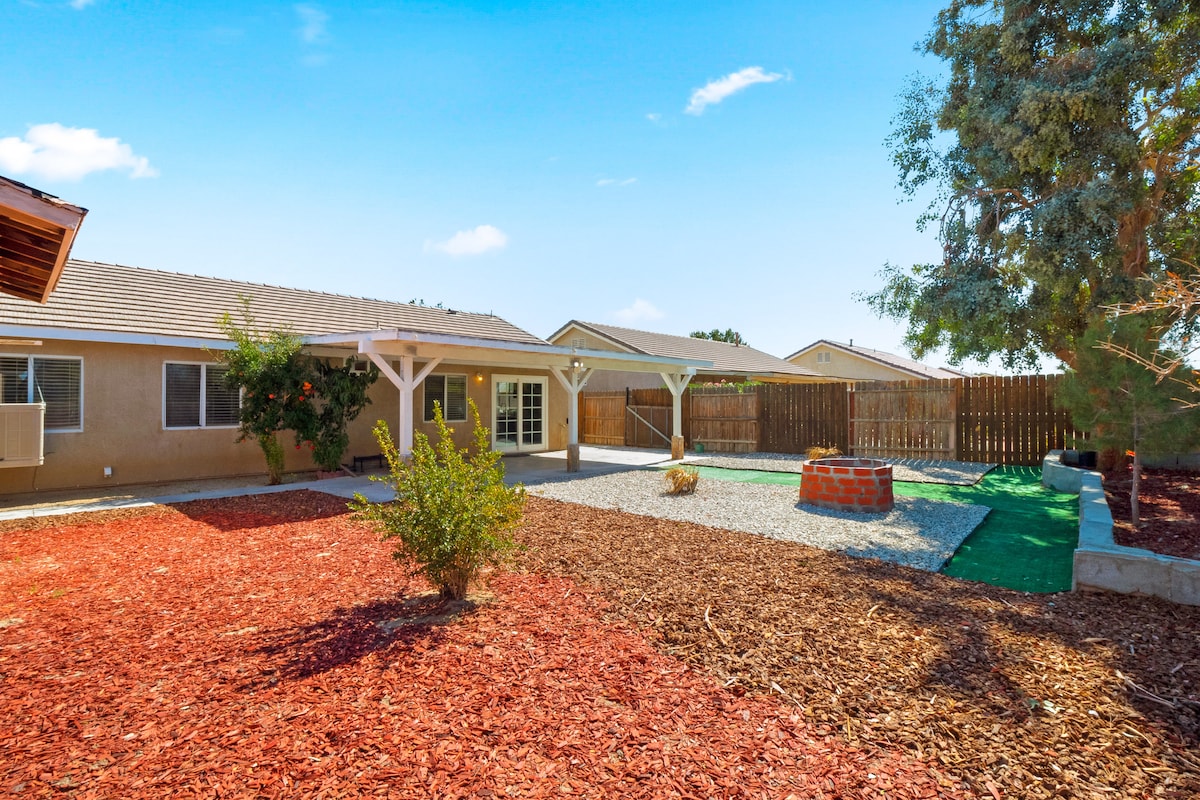 The backyard features a spacious area with gravel and mulch landscaping. A brick fire pit is positioned near the center, surrounded by pathways and mulched sections. The home’s exterior is visible in the background, with covered patios providing shaded spots for relaxation.