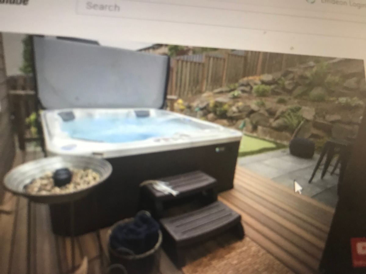 A hot tub is featured on a wooden deck, surrounded by natural stone landscaping and greenery. There are two small steps leading to the hot tub, and a metal container filled with pebbles sits nearby, adding to the outdoor ambiance.