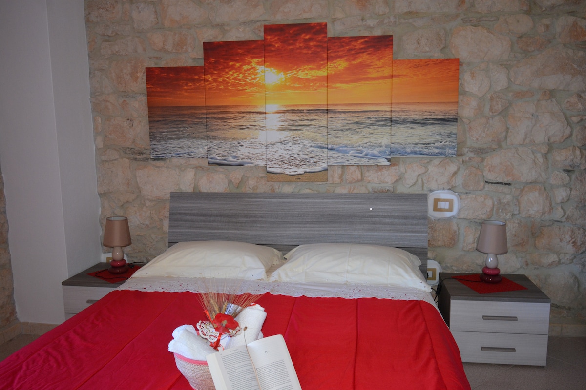 A well-appointed bedroom features a spacious bed adorned with a red coverlet. Soft bedside lamps sit on either side, complementing the natural stone wall in the background. A vibrant sunset artwork is displayed above the headboard, enhancing the calming atmosphere.