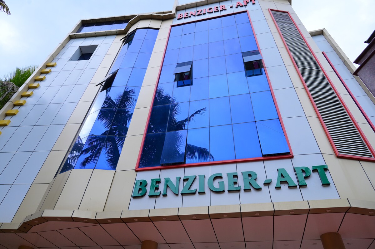 The exterior of Benziger Apartment is showcased, featuring a modern architectural design with reflective glass panels. The building name, 'BENZIGER APT,' is prominently displayed in green against the gold-toned facade, framed by tropical palm trees partially visible in the background.