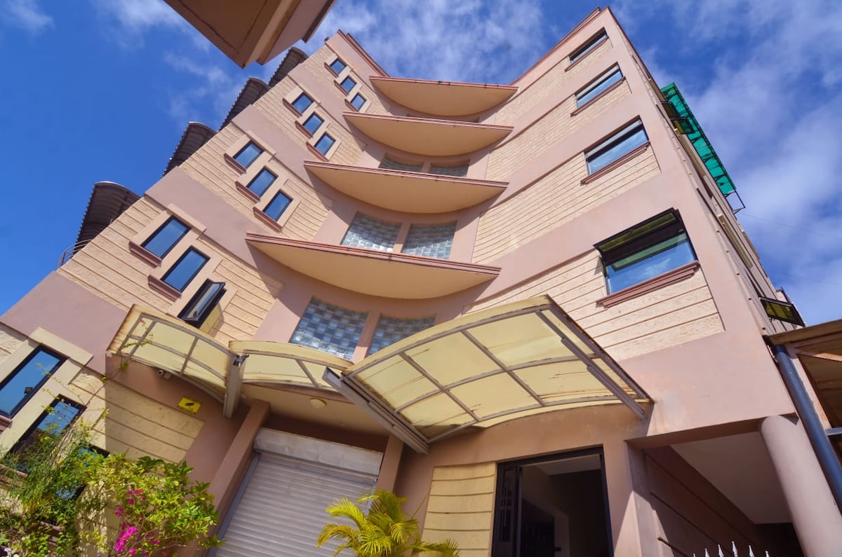The exterior of a modern five-story building is presented, featuring a unique architectural design with curved balconies. Large windows allow natural light to fill the interiors. A covered entrance and landscaped greenery enhance the welcoming appearance of the property.