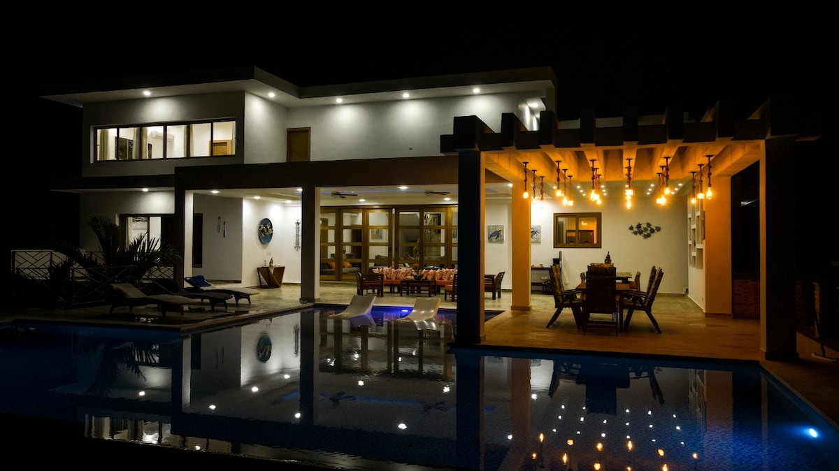 The villa is illuminated at night, highlighting its modern architecture. A large outdoor dining area features overhead lights and a spacious table. The reflection of the structure can be seen in the pool, which adds to the ambiance of the tranquil setting.