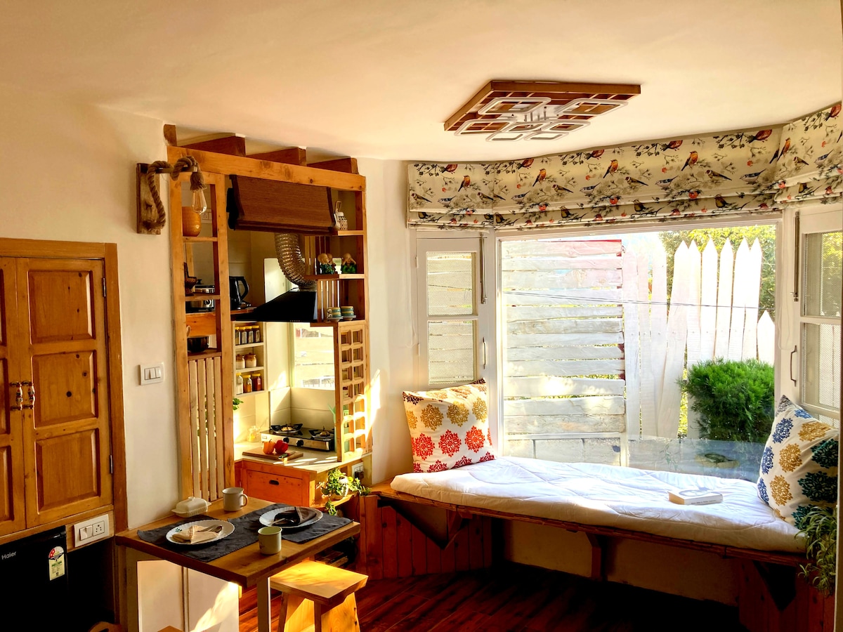 A cozy studio interior features a charming seating nook with plush cushions and a sunny window view. A wooden table is set for meals, complementing the rustic style of the cedar wood furnishings. Natural light brightens the space, enhancing the inviting atmosphere.