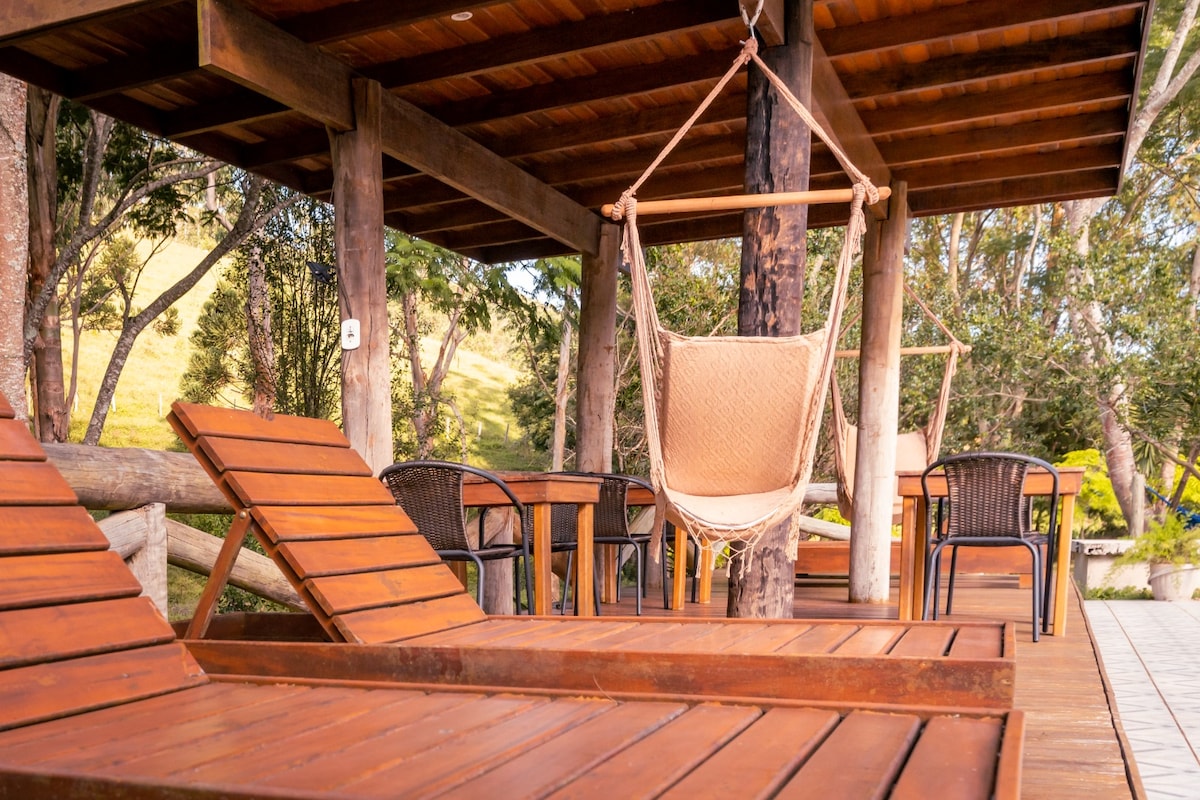 A shaded outdoor deck features wooden lounge chairs and a hammock, complemented by black metal tables and chairs. Surrounding greenery creates a natural backdrop, inviting relaxation in a tranquil setting.