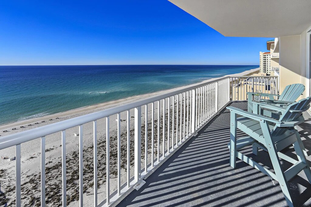 A private balcony offers expansive views of the coastline, with soft waves gently lapping against the sandy beach. Two light blue chairs are positioned to provide a comfortable spot for relaxation while enjoying the ocean scenery under a clear blue sky.