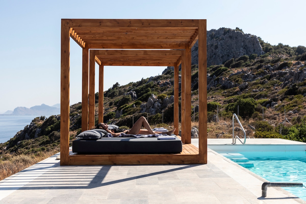 An outdoor lounge area features a wooden pergola, offering shade and a relaxed atmosphere. A person reclines on a spacious sunbed, overlooking a private pool and scenic hillside. The surrounding landscape displays rocky terrain and greenery, blending seamlessly with the deep blue water.