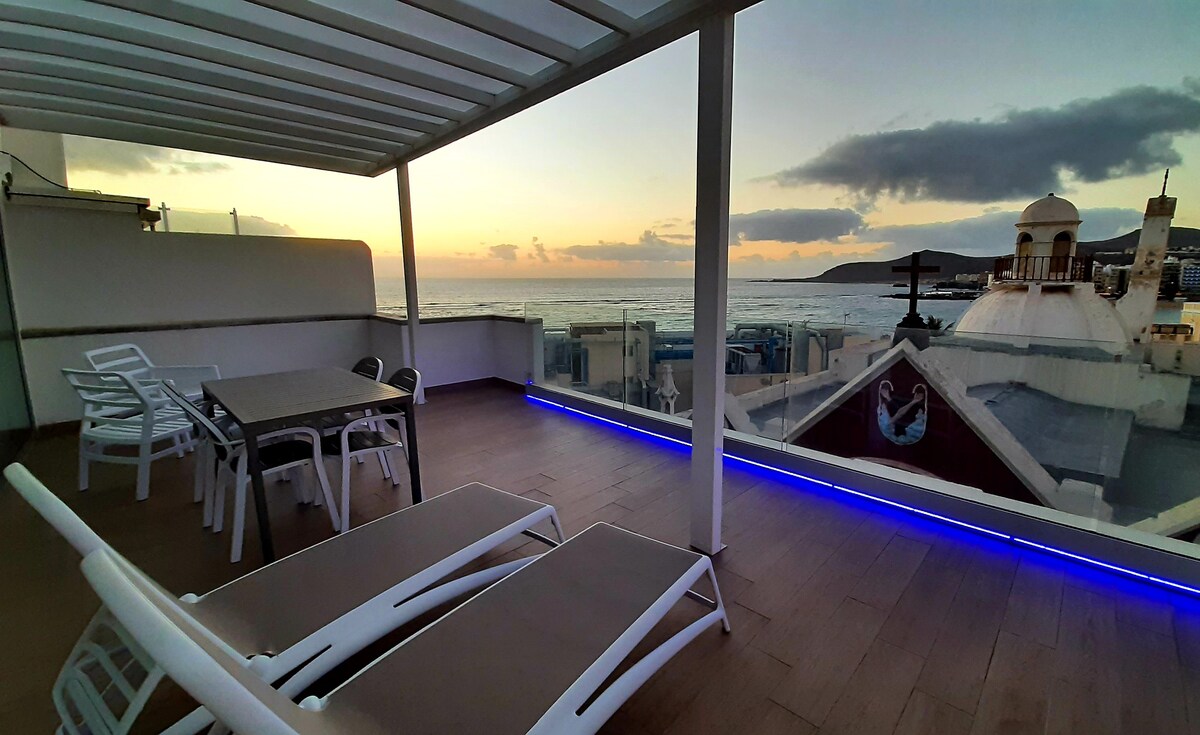 A spacious terrace offers a panoramic view of the sea during sunset. Comfortable lounge chairs are positioned for relaxation, alongside a dining table set under a shaded area. The space is illuminated with soft blue lighting, enhancing the evening ambiance.
