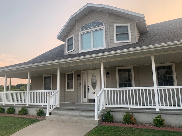 A welcoming facade of a large, two-story house is presented, featuring a centered front door with decorative glass and an arch window above. The porch, bordered by white railings, is accompanied by neatly arranged shrubs and a stone pathway leading to the entrance.