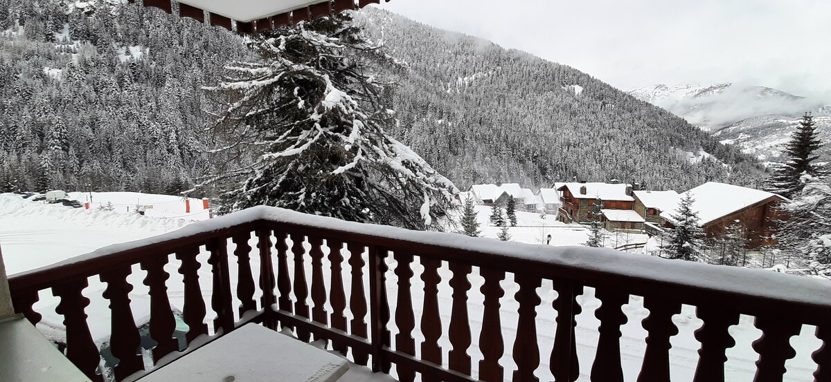 A snow-covered landscape is visible from a balcony, featuring a view of mountains blanketed in white. The balcony railing exhibits a traditional wooden design, while a few rustic buildings can be seen in the distance, surrounded by a thick layer of snow and evergreen trees.