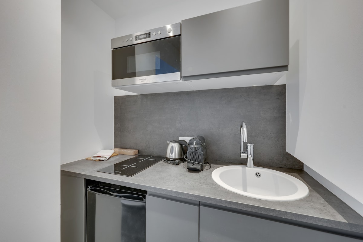 A compact kitchen area features modern, streamlined cabinetry and stainless steel appliances. An induction cooktop is bordered by a stainless steel microwave above, while a small refrigerator is tucked beneath the countertop. A round sink is positioned adjacent to the appliances, contributing to the utilitarian design.