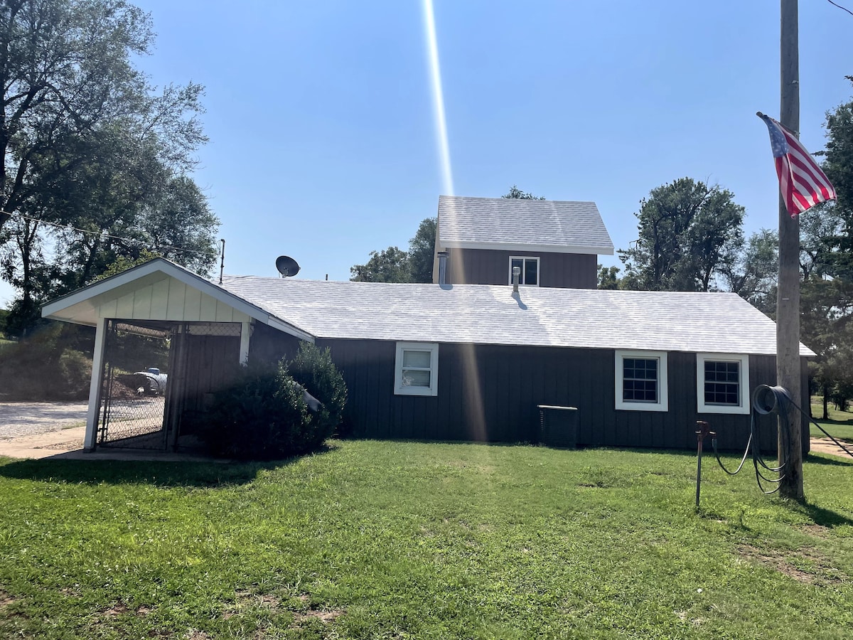 The exterior of the guesthouse is presented with a light-colored roof and dark siding. A covered porch is visible on one side, while a flagpole displaying the American flag stands nearby. Surrounding green grass and trees add to the inviting environment.