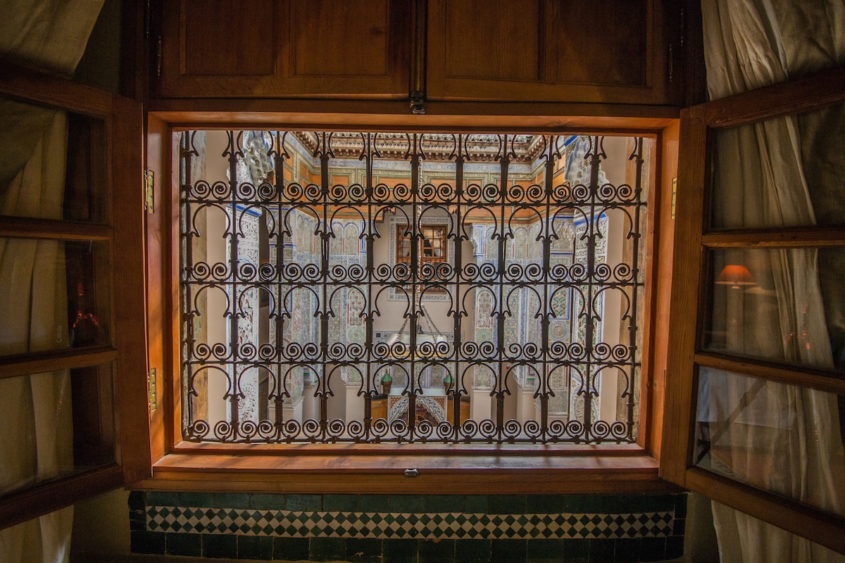 A wooden window frame presents an intricate wrought iron grille, looking out into a courtyard. Sunlight filters through, casting soft shadows on the walls. Colorful tiles adorn the lower part of the window, adding to the architectural charm.