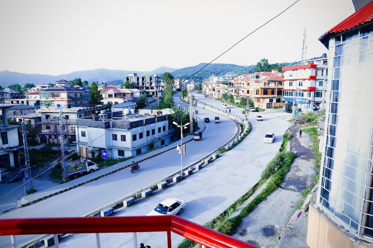 A view from the balcony captures the winding road lined with buildings and trees. The scene features a mix of residential and commercial structures, with vehicles visible on the road. The backdrop showcases rolling hills under a clear sky.