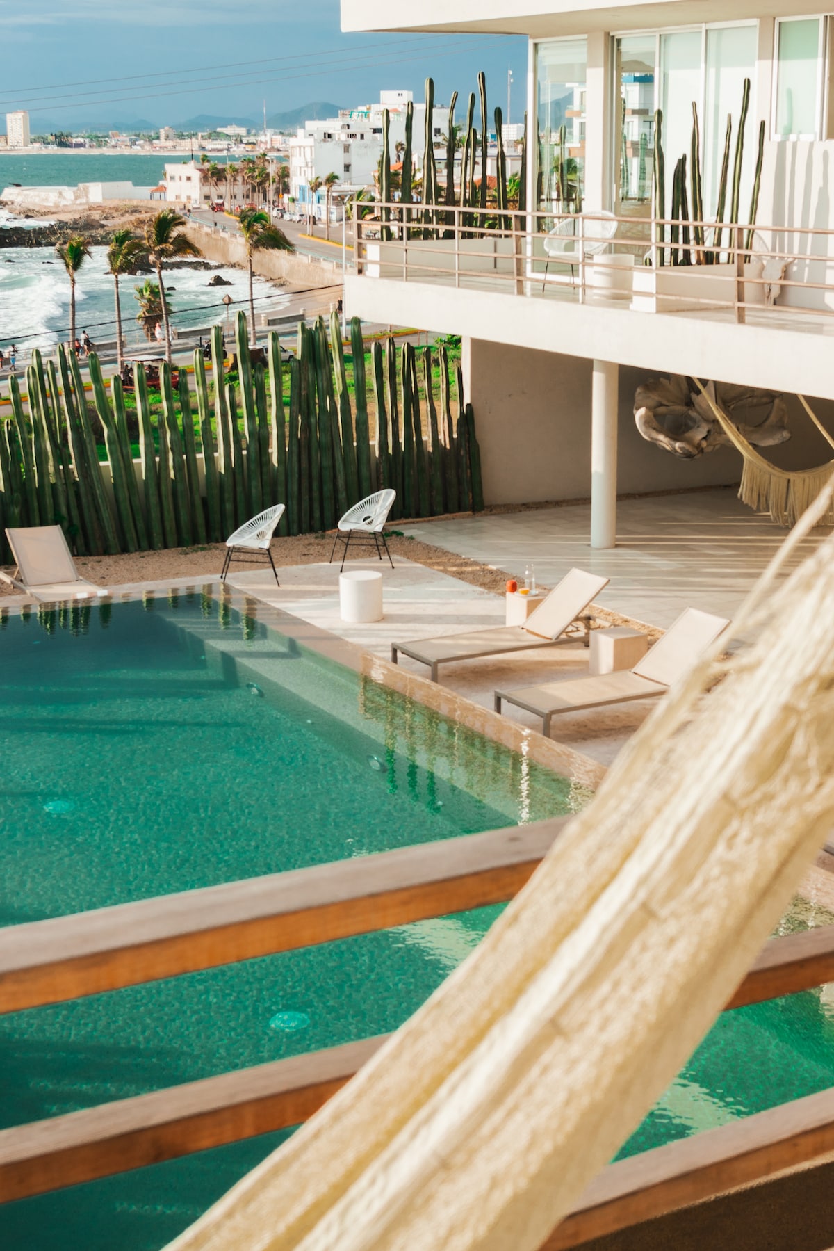 A serene pool area is visible, surrounded by lounge chairs and complemented by a hammock. The ocean can be seen in the background, along with palm trees and cacti, creating a relaxed coastal ambiance. A modern building with large windows provides an expansive view.