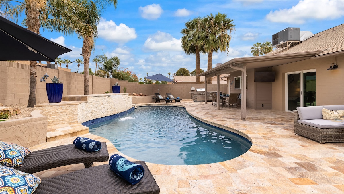 A resort-style backyard features a sparkling pool surrounded by natural stone patios. Comfortable lounge chairs are positioned by the water, and large palm trees add a tropical touch. A shaded area with an umbrella and an inviting sofa complete the outdoor living space.