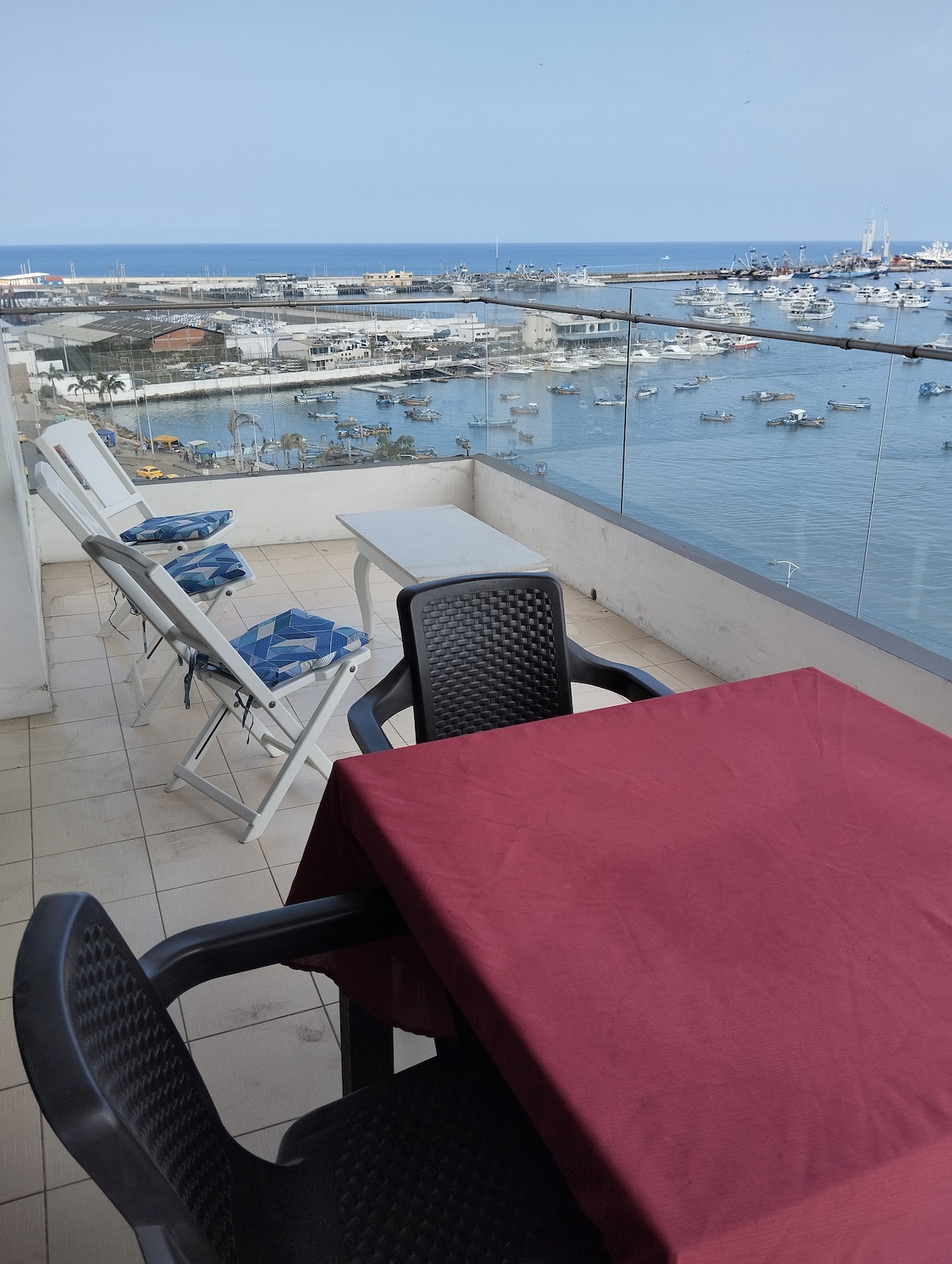 A balcony offers views of the marina, with the ocean visible in the background. A dining table with a maroon tablecloth sits beside two lounge chairs. The space is well-lit and provides an inviting area to relax while enjoying the coastal scenery.
