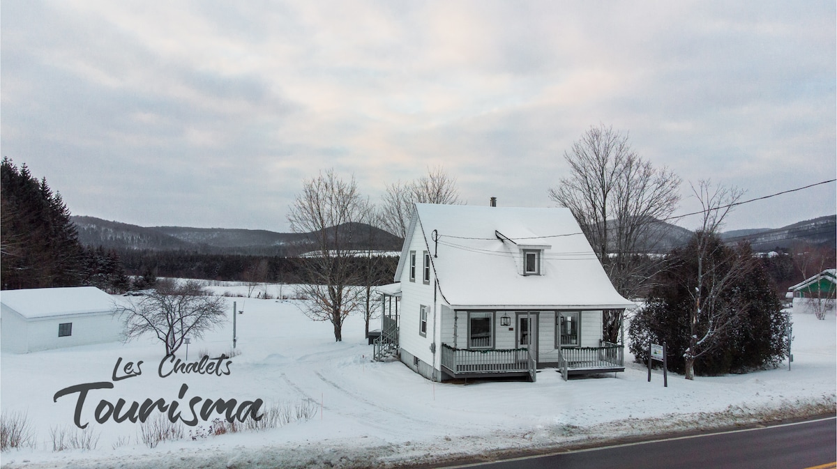 A charming two-story house is shown, surrounded by a snowy landscape. The building features a covered porch and multiple windows. Nearby, open fields and distant hills are visible, covered in a blanket of white. The scene suggests a tranquil winter setting.