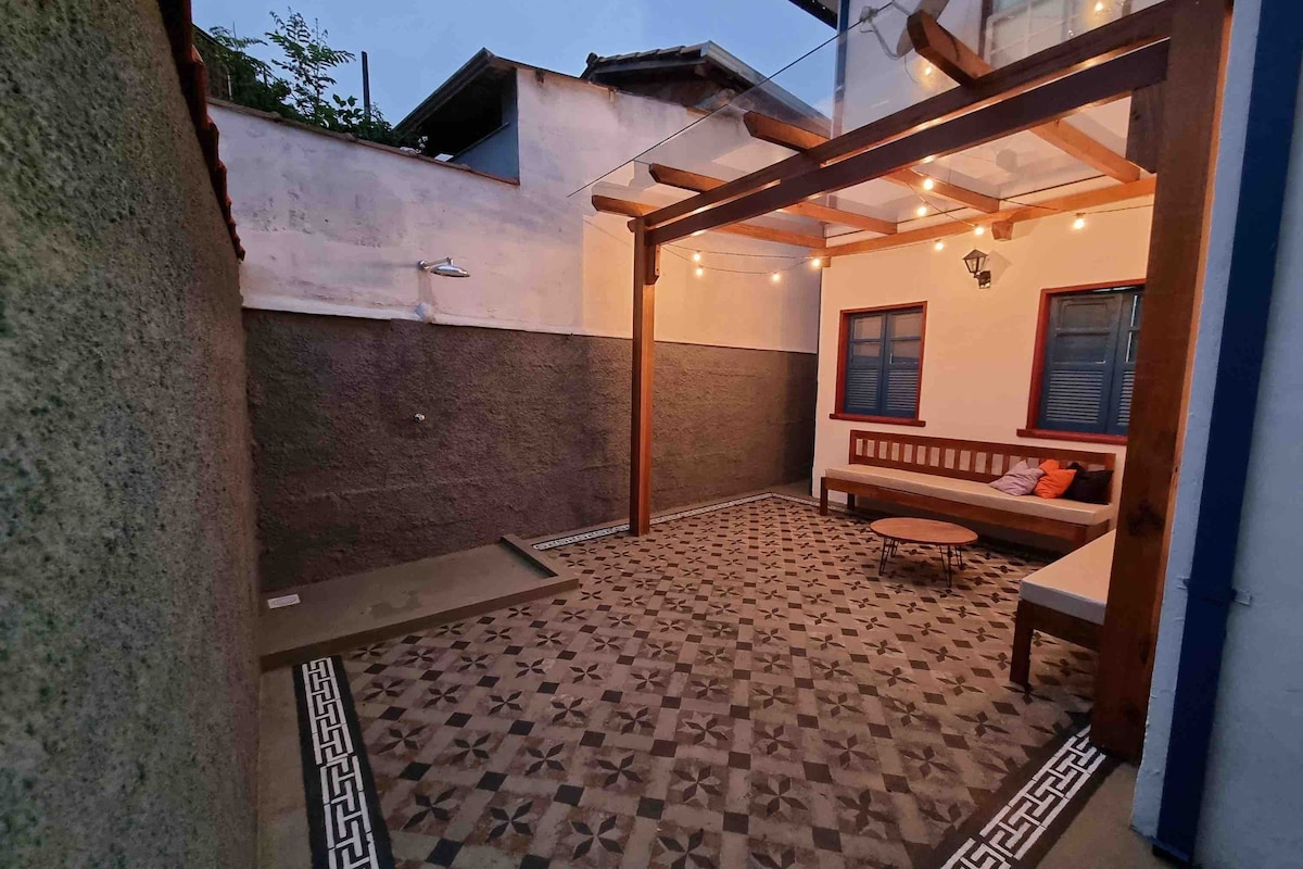 An outdoor area features geometric patterned tile flooring, complemented by wooden beams supporting a glass roof. Soft lighting illuminates a cozy seating space with a fabric-covered bench and a round coffee table, creating an inviting atmosphere for relaxation or gatherings.