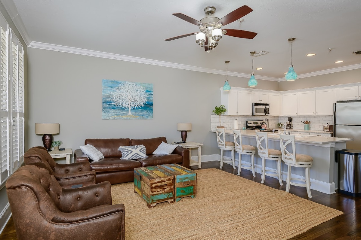 A spacious living area features a comfortable brown leather sofa and two armchairs, arranged around a rustic coffee table. Bright pendant lights illuminate the open kitchen area, which is equipped with modern appliances. Light-colored walls and a large rug contribute to a welcoming ambiance.