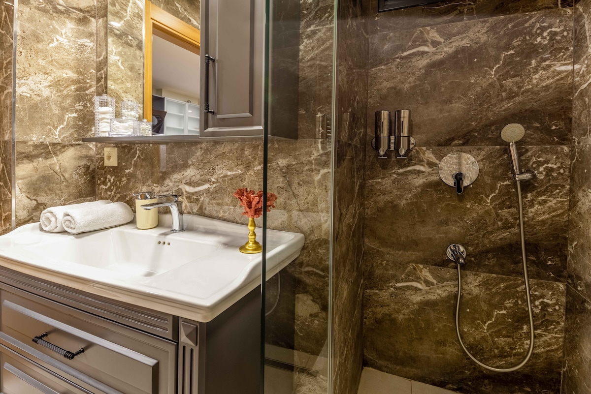 A stylish bathroom features elegant stone walls, with a modern shower area and a sleek countertop. High-quality toiletries are neatly arranged beside the sink, complemented by a decorative item. Ample lighting enhances the contemporary design.