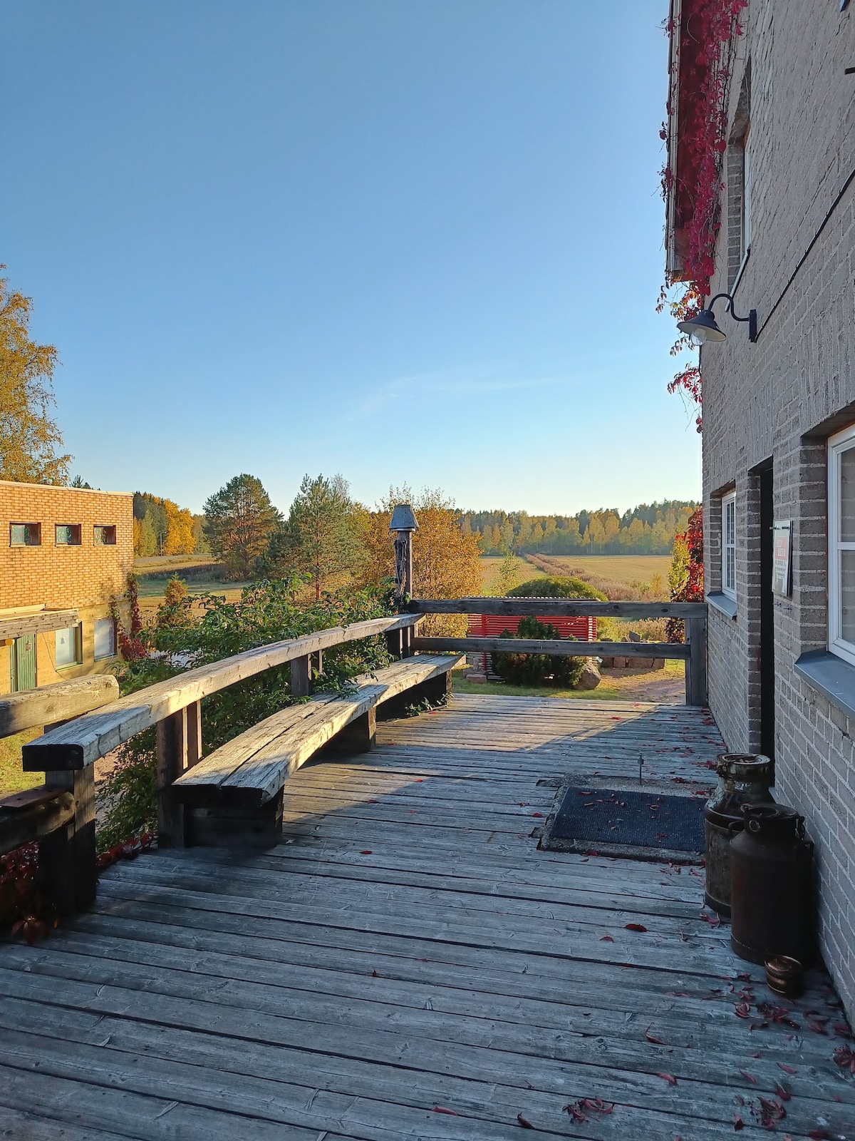A wooden walkway is shown leading to an expansive view of neighboring fields and trees, framed by autumn foliage. The bright blue sky complements the vibrant colors of the landscape, enhancing the outdoor space's inviting atmosphere.