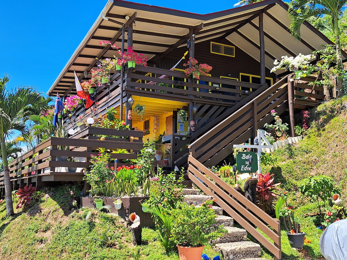 The house features a wooden exterior embraced by lush greenery and colorful flowers. A series of steps lead to a welcoming entrance. Several potted plants are arranged along the path, while a vibrant sign is displayed near the base, contributing to the inviting atmosphere.