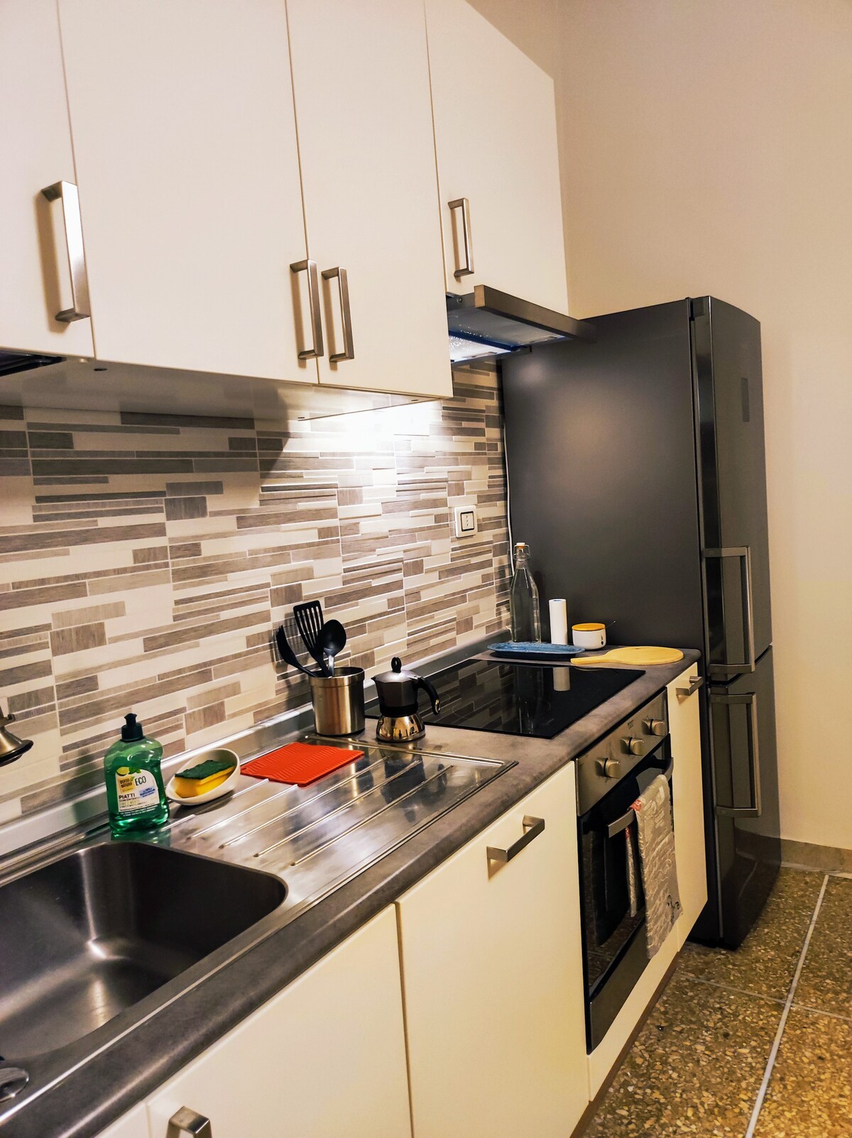 A modern kitchen is equipped with sleek cabinetry and a stone countertop. Stainless steel appliances, including a refrigerator and oven, complement the space. The mosaic backsplash adds visual interest, while the arrangement of utensils and kitchenware suggests functionality.