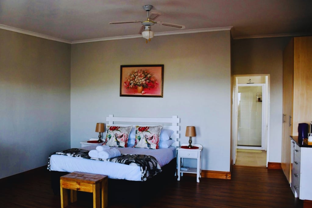 A spacious room features a bed with floral-patterned pillows, accompanied by bedside tables and lamps. A wooden stool sits at the foot of the bed. Light-colored cabinetry is visible, with a doorway leading to a bathroom with a shower.