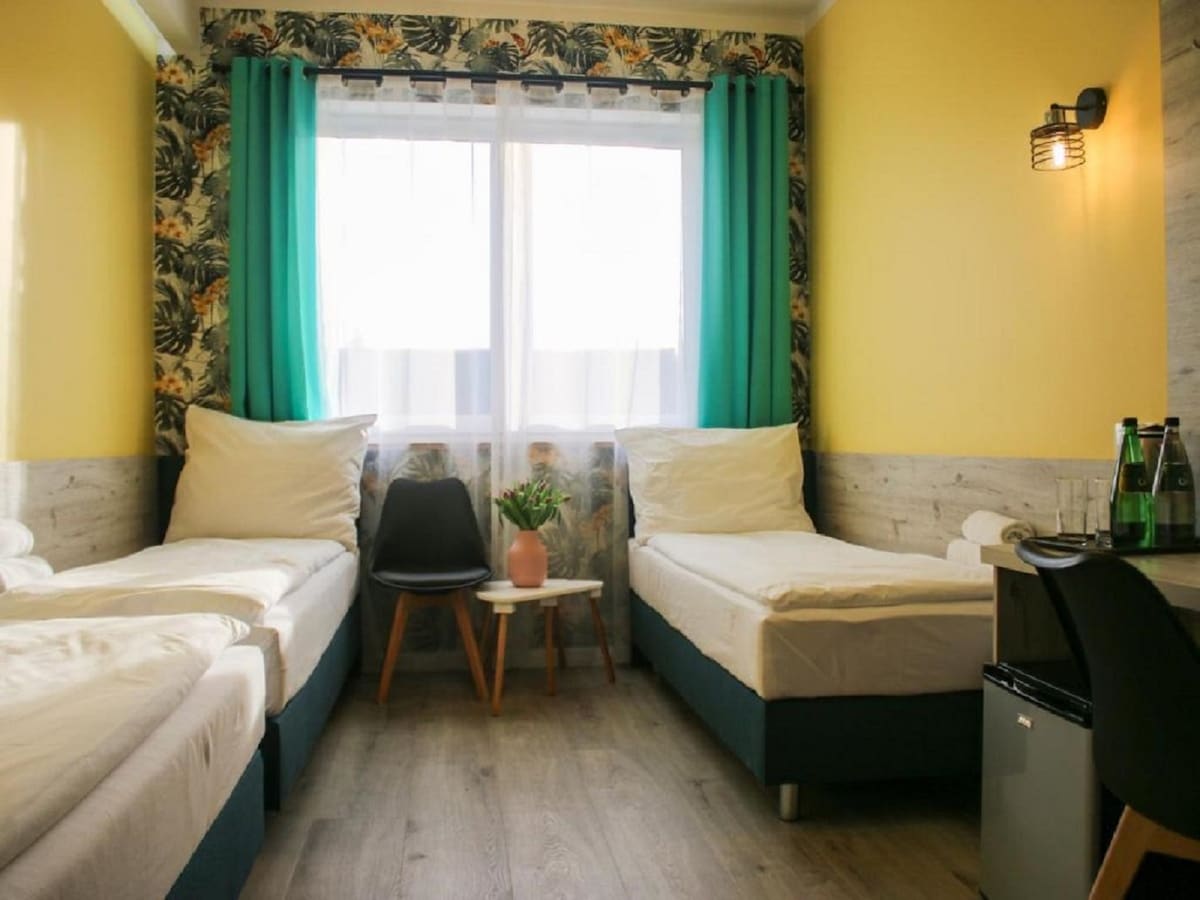 The room features three individual beds, arranged to maximize space, alongside a small table and two chairs. Large windows are dressed with teal curtains, allowing natural light to brighten the area. A refrigerator is positioned near the wall, enhancing functionality.