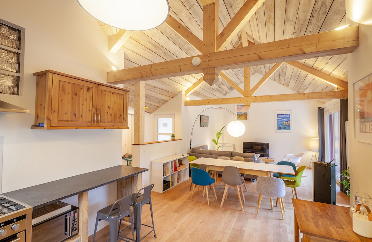 An open living area features exposed wooden beams and warm tones. A dining table is surrounded by colorful chairs, while a cozy seating arrangement is positioned nearby. A large window allows natural light to brighten the space, and a modern lamp complements the design.