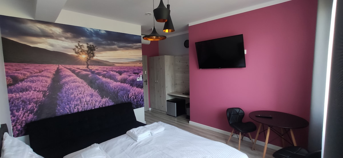 A spacious room features a large double bed with white linens, complemented by a vibrant lavender-themed wall mural. A black TV is mounted on the pink wall, and a small dining table with two chairs is visible next to a wooden desk.