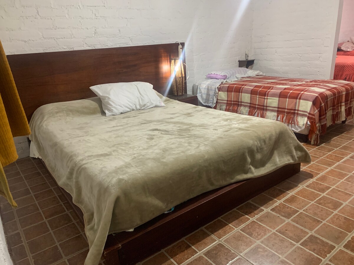A spacious sleeping area features a large bed covered with a cozy beige blanket, and a smaller bed dressed in a plaid blanket. Soft lighting highlights the room's neutral color scheme and textured walls.