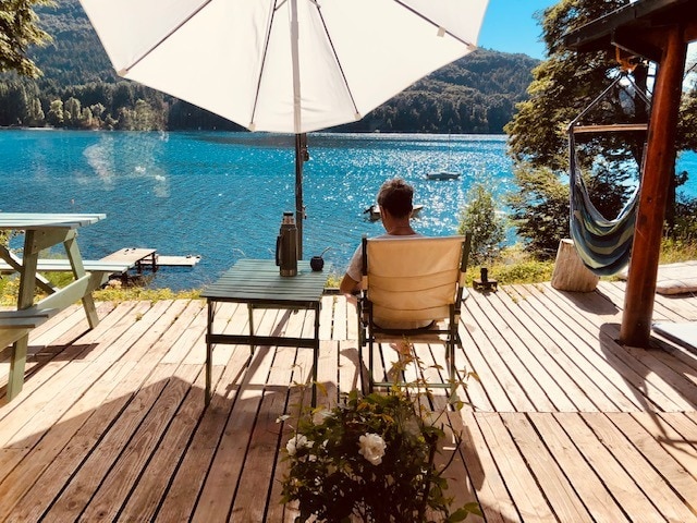 A serene outdoor seating area features wooden deck boards and a sunshade umbrella. A guest is seated in a chair, facing a calm lake with glistening water. Nearby, a relaxing hammock is visible, surrounded by lush greenery.