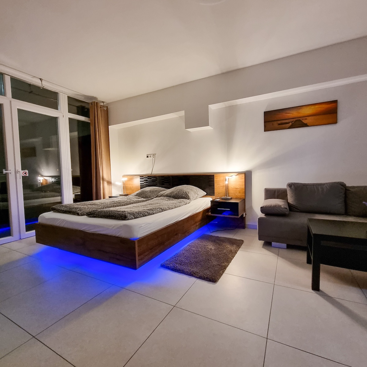 A modern sleeping area features a spacious bed with soft bedding, accentuated by subtle blue lighting. A cozy seating area includes a gray couch and a small coffee table. Large glass doors allow natural light and views of the outside.