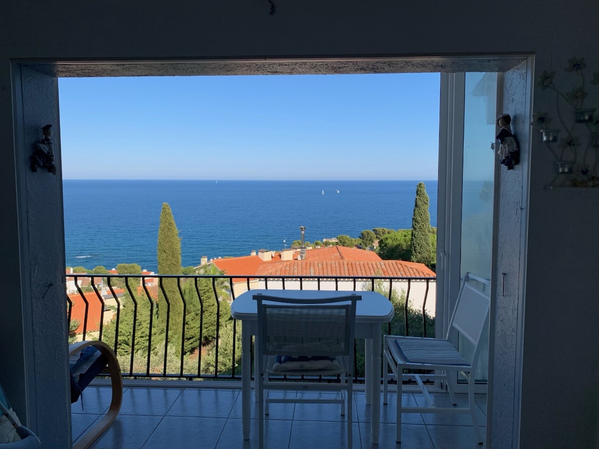A serene view of the ocean is framed by an open doorway. A small table and two chairs are positioned on the balcony, overlooking the tranquil water and lush greenery below. The clear blue sky complements the soothing coastal landscape.