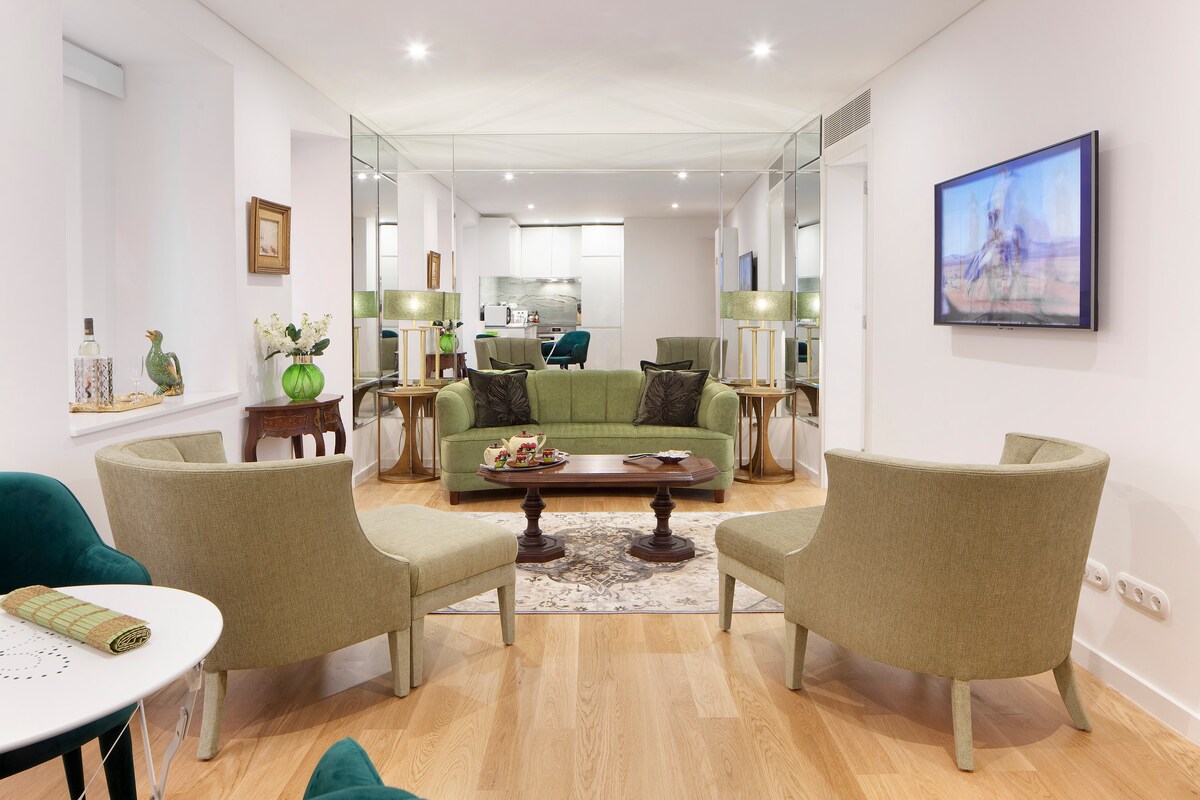 A stylish living area is arranged with two green armchairs flanking a wooden coffee table. A neutral rug is placed on the wooden floor, while a wall-mounted television is visible above the table. A modern kitchen can be seen in the background.