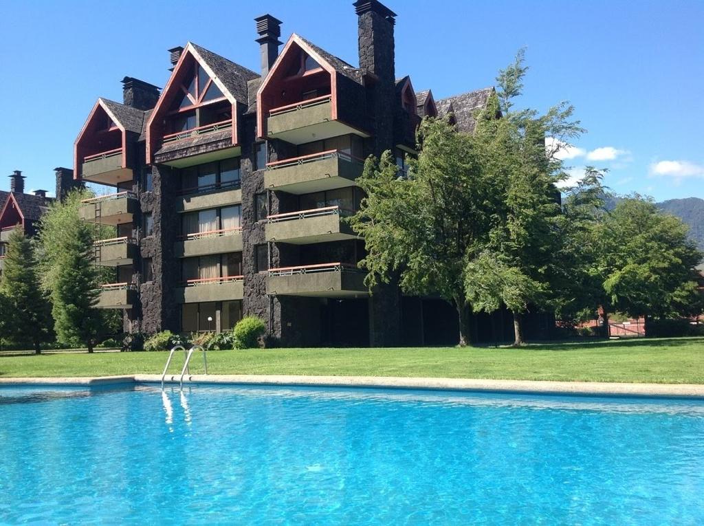 A three-story building features a mix of dark stone and wood details, with several balconies visible. A sparkling blue pool is nestled in the foreground, surrounded by lush green grass and trees, reflecting the clear blue sky.